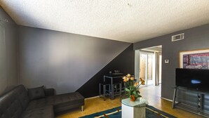 Een moderne woonkamer met een grijze bank, een bijzettafeltje met een bloevas, een flatscreen televisie en een trap die naar een andere kamer leidt.