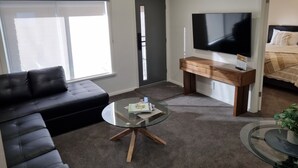 A modern living room with a black leather sofa, a glass coffee table, a wooden TV stand with a flat-screen TV, and a bed with a striped blanket. - Golden Heritage Accommodation Rutherglen (Rutherglen)