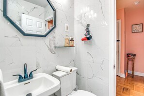Un baño con lavamanos blanco, inodoro y espejo. Hay artículos de aseo en la pared y un secador de pelo.