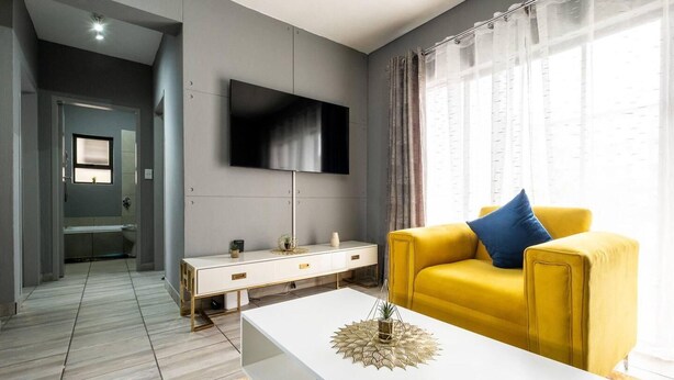 A modern living room with a yellow sofa, a white coffee table, and a flat-screen TV mounted on the wall. - Sleeps4-pool-hottub-petfriendly-garden (Johannesburg)
