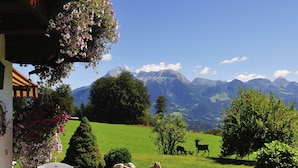 A patio with a table and chairs, a stone pathway, and a view of a green field with deer and mountains in the background. - Fewo Nr. 2 Lattenberg für 2-3 Pers., 1 Schlafzimmer, Wohnraum mit Küchenzeile, Balkon (Bischofswiesen)
