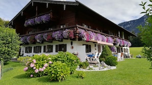 A traditional wooden chalet with balconies adorned with flowers, a well-maintained garden, and a view of mountains in the background. - Fewo Nr. 2 Lattenberg für 2-3 Pers., 1 Schlafzimmer, Wohnraum mit Küchenzeile, Balkon (Bischofswiesen)