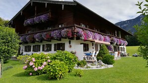 A traditional wooden chalet with balconies adorned with flowers, a well-maintained garden, and a view of mountains in the background.