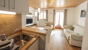 A modern kitchen with a wooden countertop, stainless steel sink, and a built-in oven. There is a dining area with a white table and chairs, and a living room with a sofa and a window with curtains.