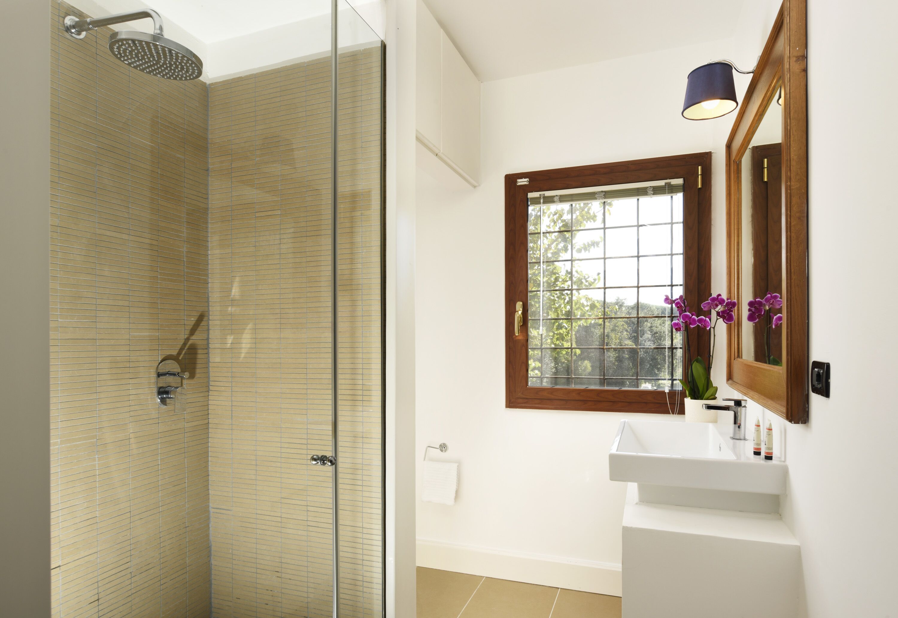 A bathroom with a glass shower enclosure, a white sink, a wooden-framed window, and a mirror with a purple orchid.