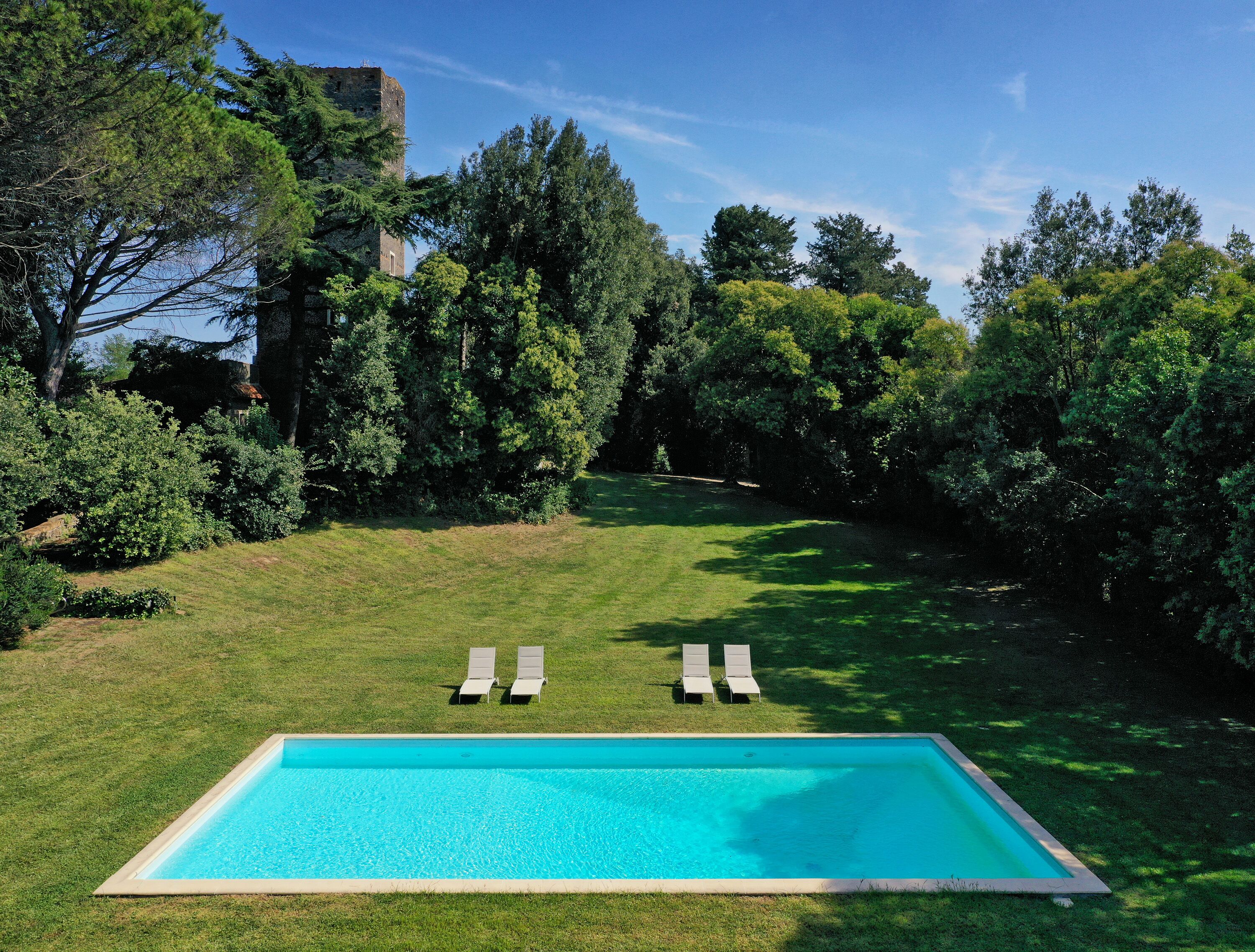 A rectangular swimming pool with two lounge chairs facing it, surrounded by a well-maintained lawn and dense trees.