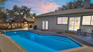 A rectangular swimming pool with a tiled edge, surrounded by a gravel area. There are outdoor chairs and a table near the pool, and a covered patio area with umbrellas. - San Antonio Escape. Close to Lackland, SeaWorld, Fiesta Texas, and Riverwalk (San Antonio)