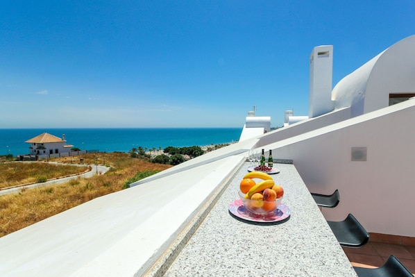 A balcony with a fruit bowl, a bottle of wine, and a view of the sea.