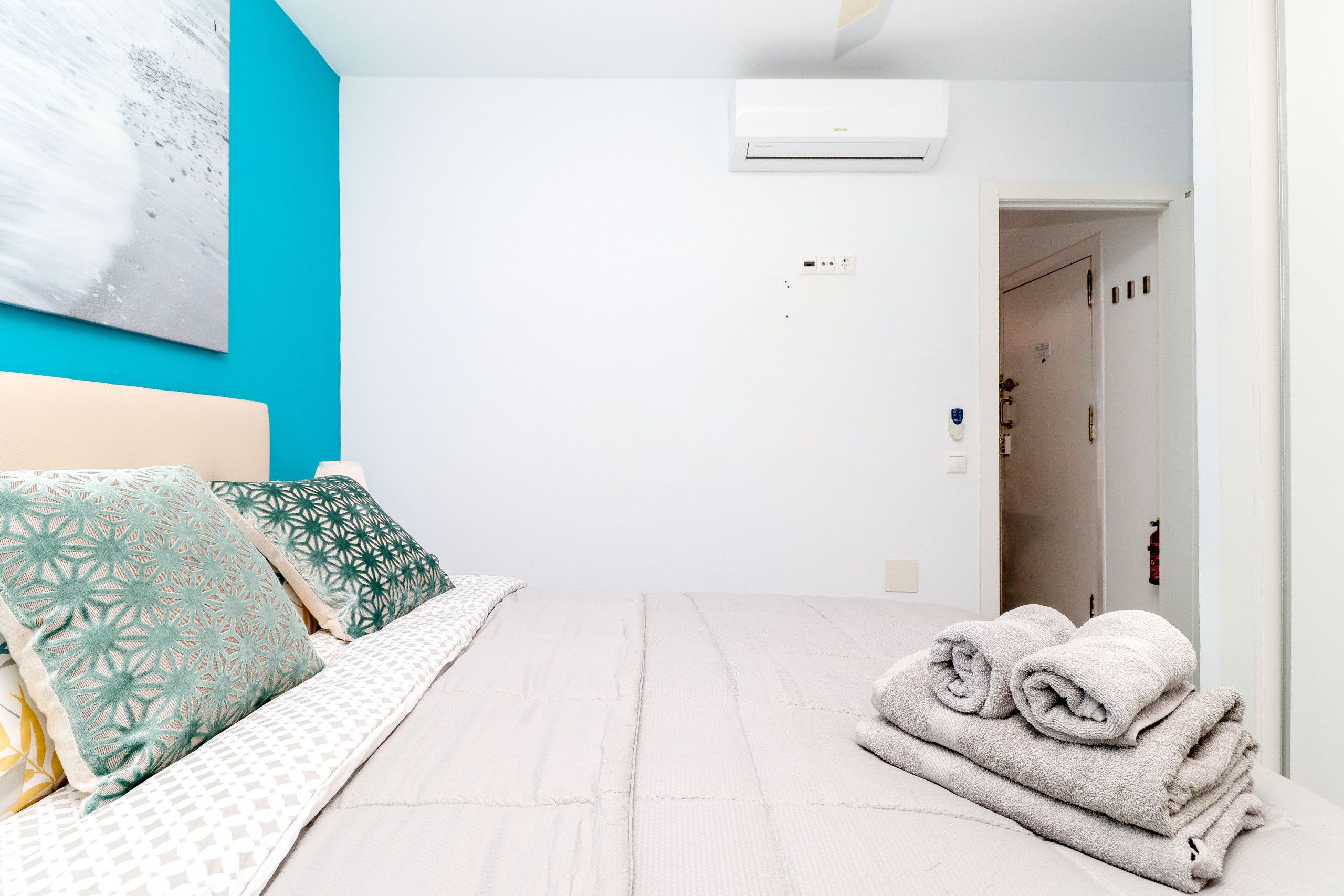 Un dormitorio con cama, almohadas, aire acondicionado de pared y una puerta que da a otra habitación.