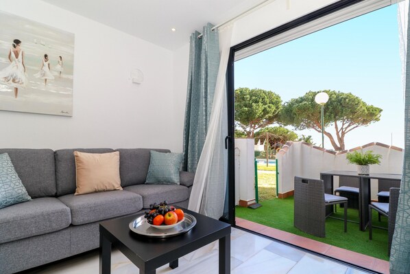 Una sala de estar moderna con un sofá gris, una mesa de centro negra con un frutero y un amplio ventanal con vistas a un jardín y árboles.