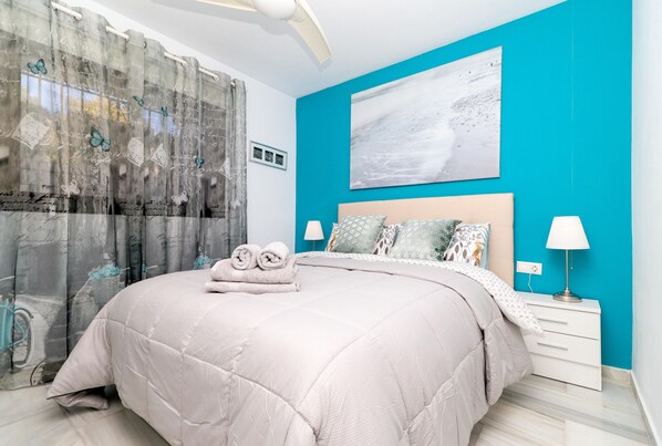 Un dormitorio con cama, mesita de noche, lámpara, ventana con cortinas y un cuadro en la pared.