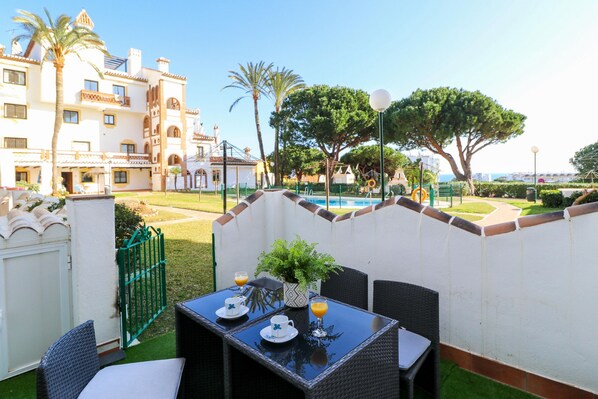 Une terrasse extérieure avec une table dressée pour deux personnes, entourée de chaises en osier, d’une plante en pot et offrant une vue sur une piscine et des bâtiments en arrière-plan.