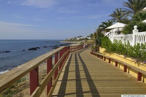 A wooden boardwalk with red railings leading to a beach, flanked by palm trees and a white building with a balcony. - Calahonda apartment - all facilities 5 mins walk (Mijas Costa)