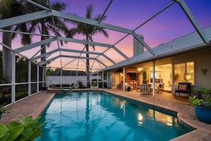 A swimming pool with a glass enclosure, surrounded by palm trees and a sunset sky. - Luxury Canal Home | Pool, Spa & Dock Near Beach (Nokomis)