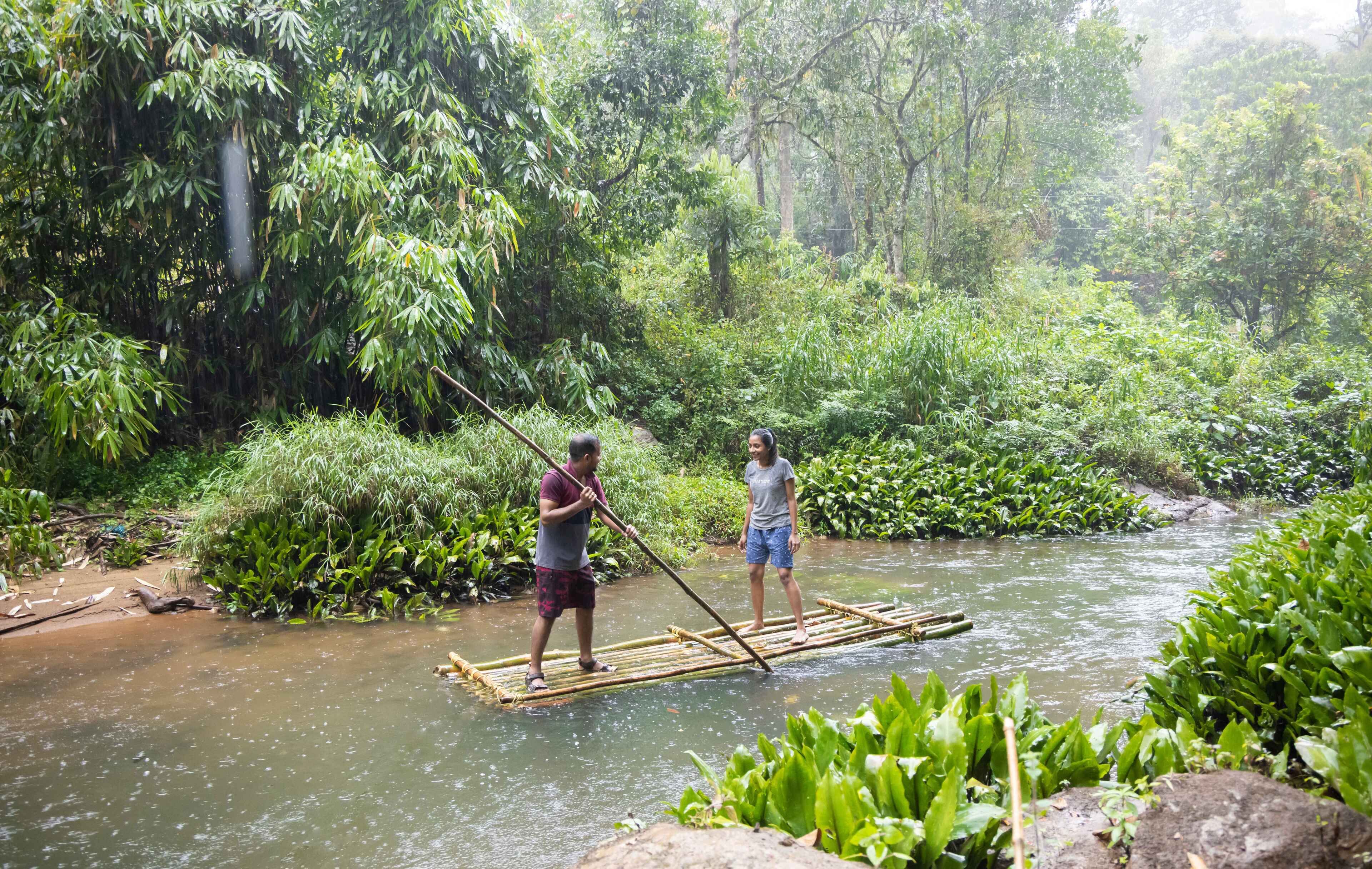 Two people standing on a bamboo raft in a river surrounded by dense greenery.