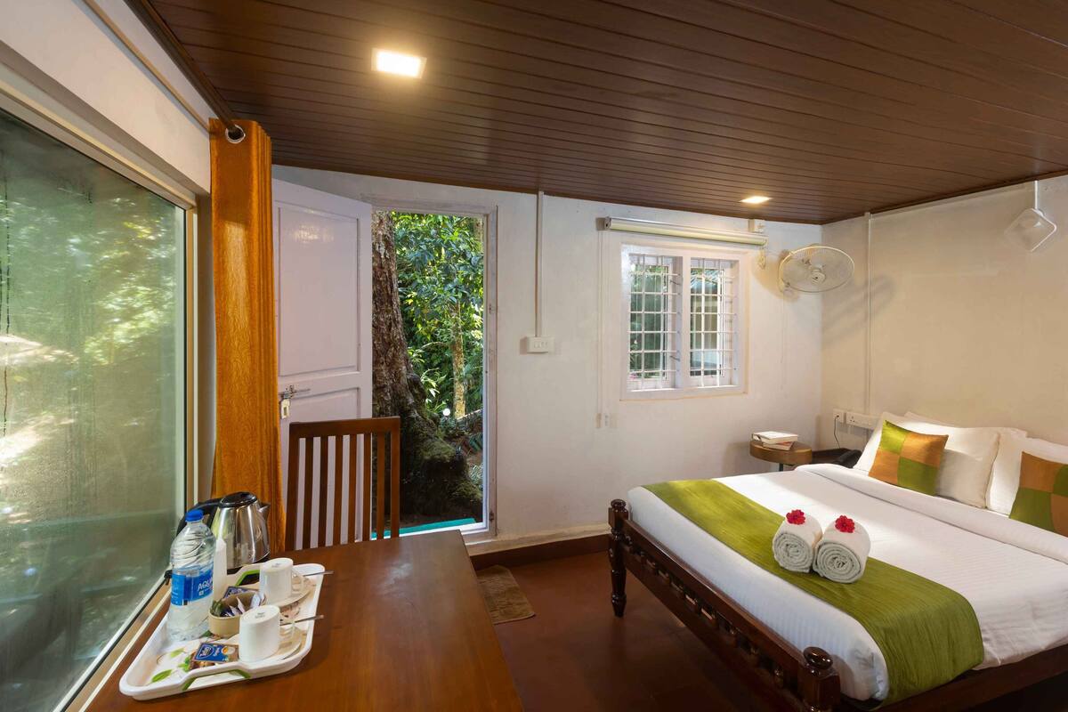 Quarto de hotel com cama, mesa com itens de café da manhã, um ventilador e vista para o verde lá fora.