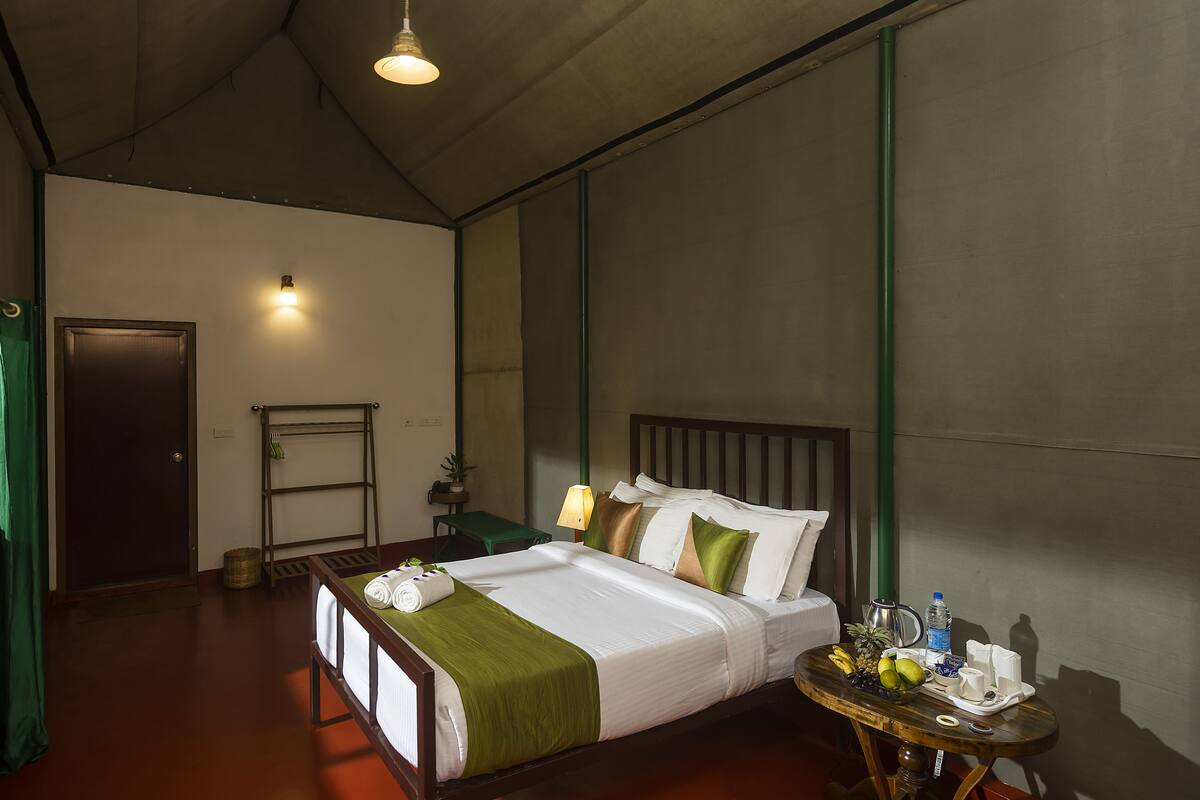 Quarto de hotel com cama, mesa de cabeceira, luminária e uma pequena mesa com frutas e uma garrafa.