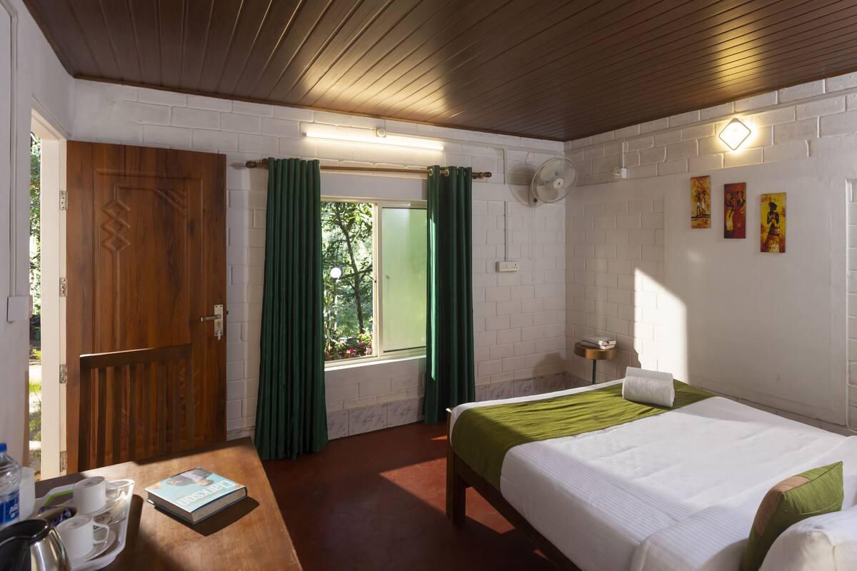 Quarto com duas camas, porta de madeira, janela com cortinas verdes, parede de tijolos brancos e uma pequena mesa com um livro e xícaras.