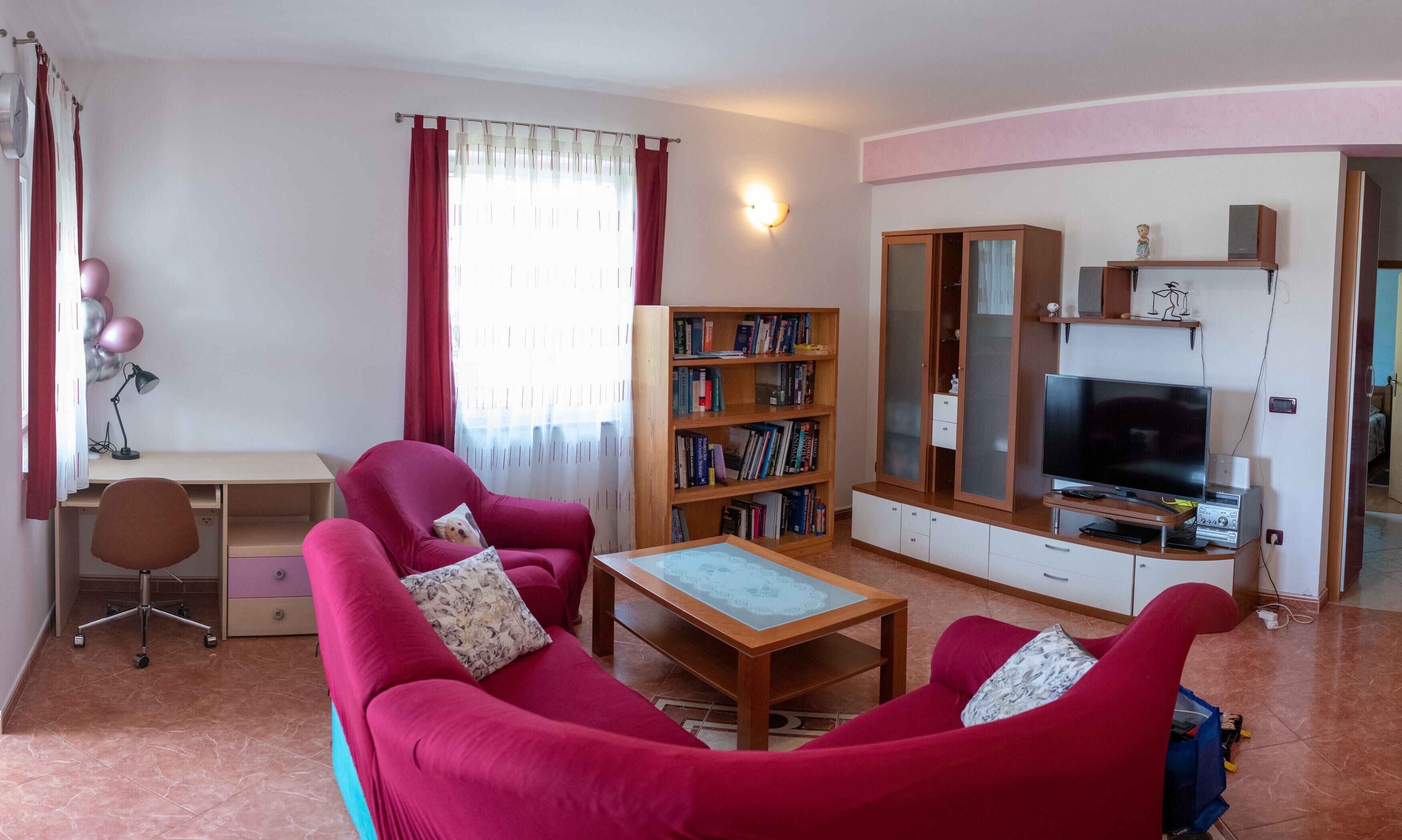 A living room with a red sofa, a wooden coffee table, a bookshelf, a TV, and a desk with a lamp.