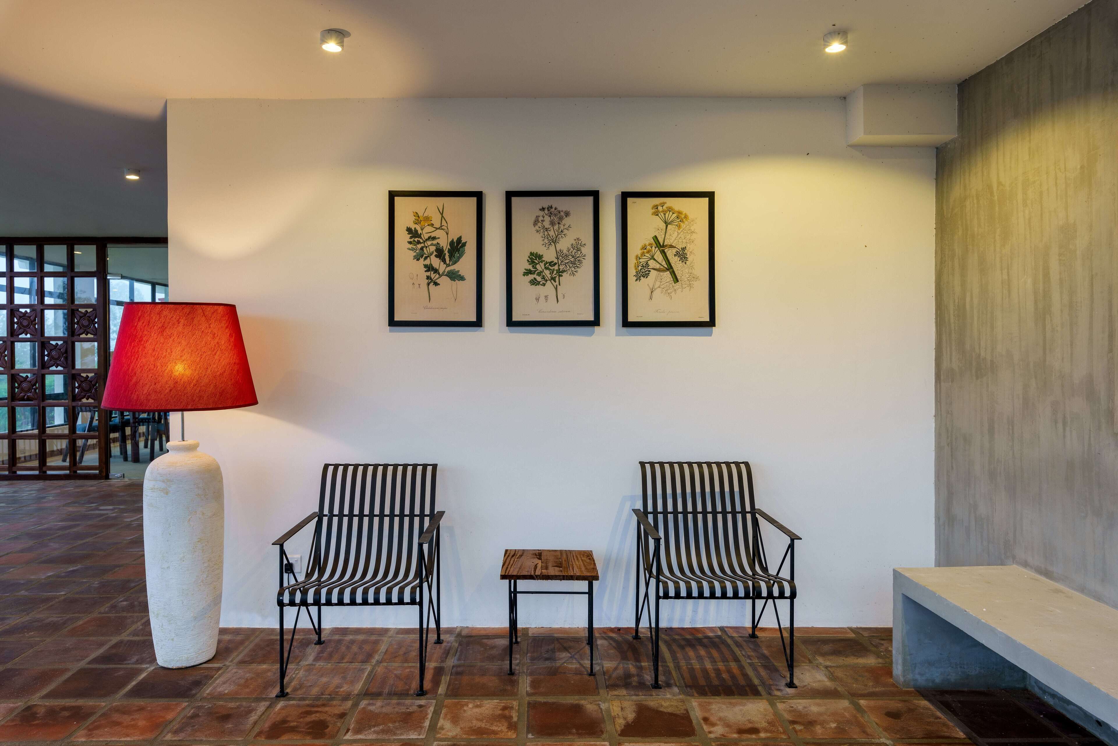 A modern lobby with a tiled floor, two striped chairs, a small table, a red lamp, and three framed botanical illustrations on the wall.
