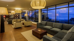 A modern hotel lobby with a large sofa, armchairs, a wooden coffee table, and large windows offering a view of the cityscape at dusk.