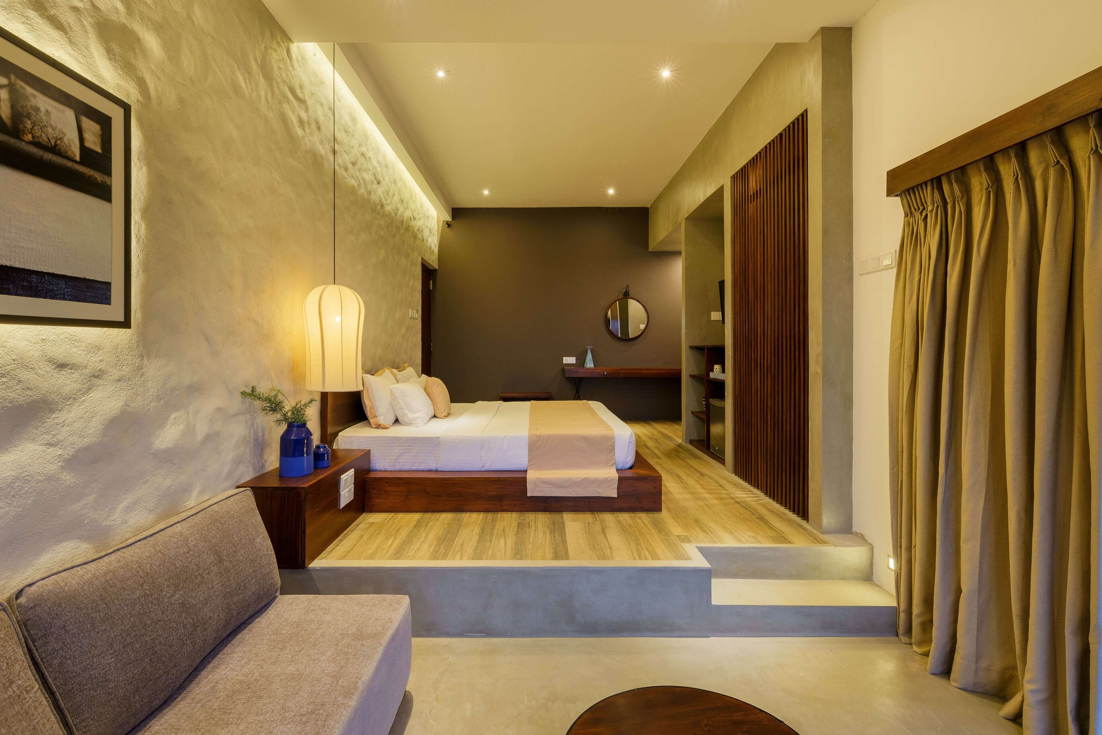A modern hotel room with a bed, a sofa, a bedside table with a lamp, a mirror, and a view of the outdoors.