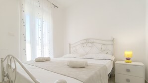 Ein ordentlich bezogenes Bett mit einem Metall-Kopfstück, ein Nachttisch mit Lampe und ein Fenster mit durchsichtigen Vorhängen.