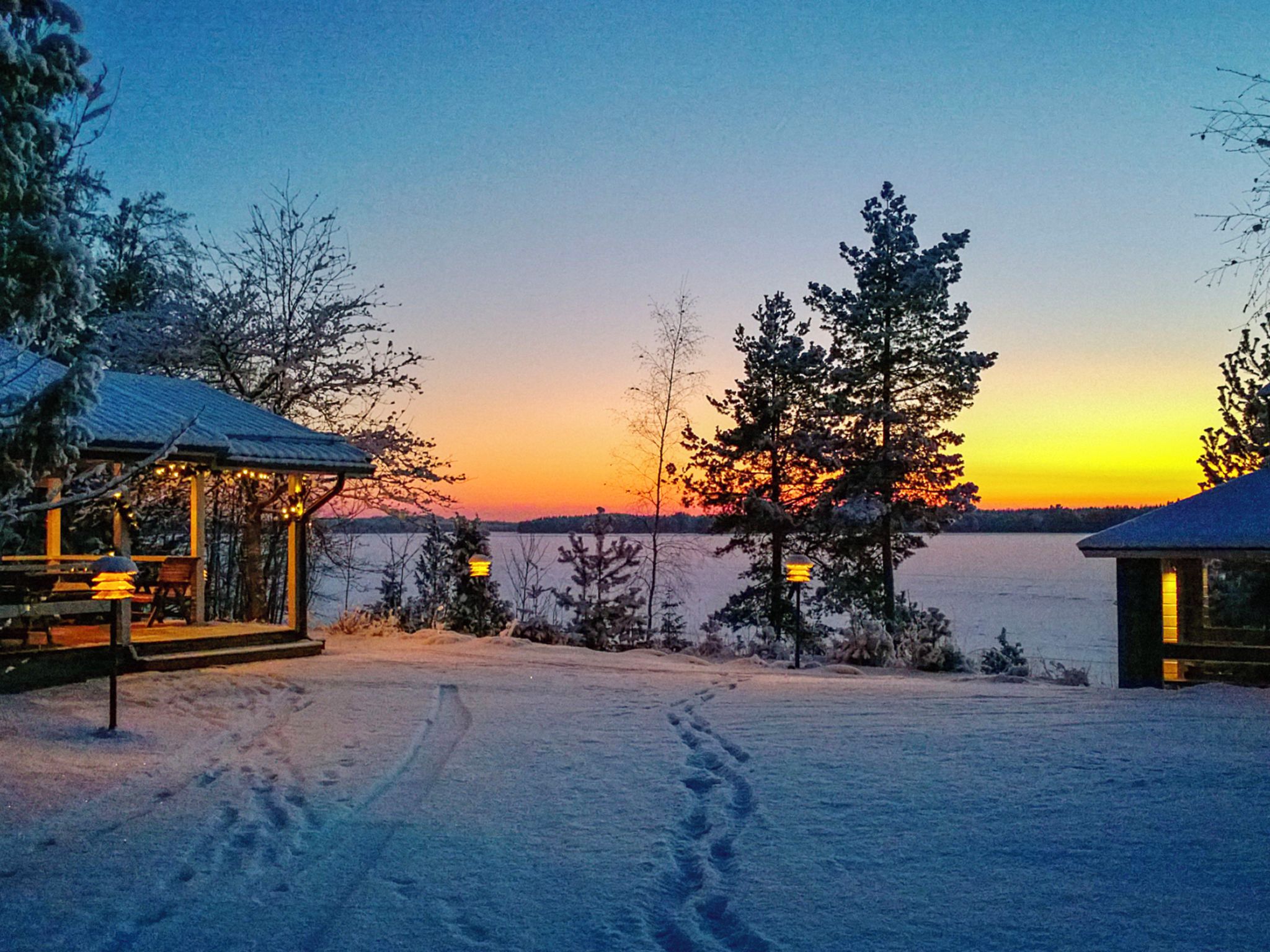 A snowy landscape with a sunset, a wooden cabin, and a clear view of the water.