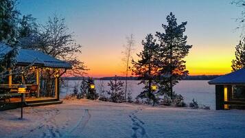 A snowy landscape with a sunset, a wooden cabin, and a clear view of the water.