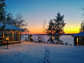Un paysage enneigé, avec un coucher de soleil, un chalet en bois et une vue dégagée sur l’eau.