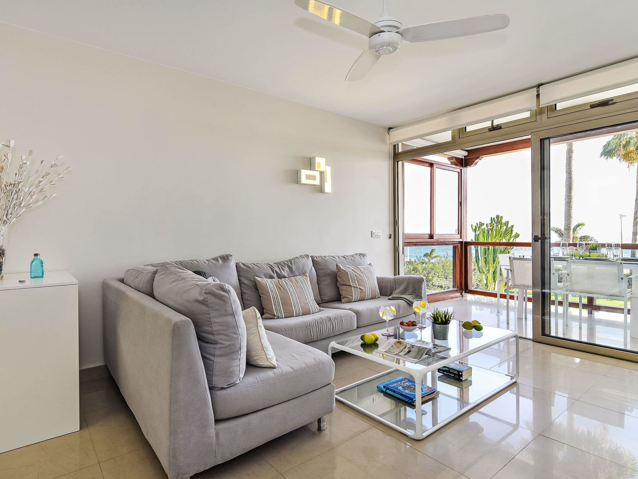 A modern living room with a grey sofa, a glass coffee table, and a ceiling fan.