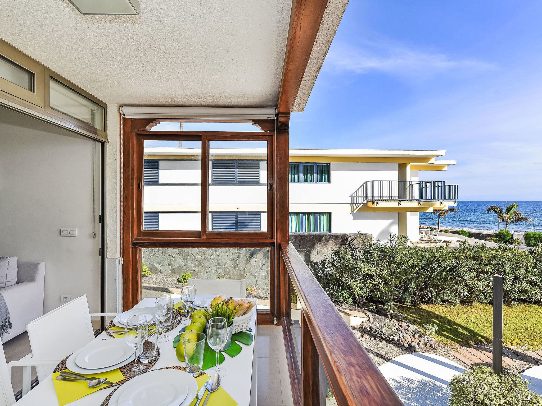 A balcony with a table set for dining, overlooking a coastal area with a building and ocean.