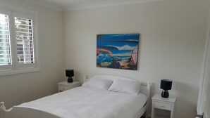 Een slaapkamer met een wit bed, twee nachtkastjes met lampen, een raam met zonwering, en een schilderij met een strandthema aan de muur.