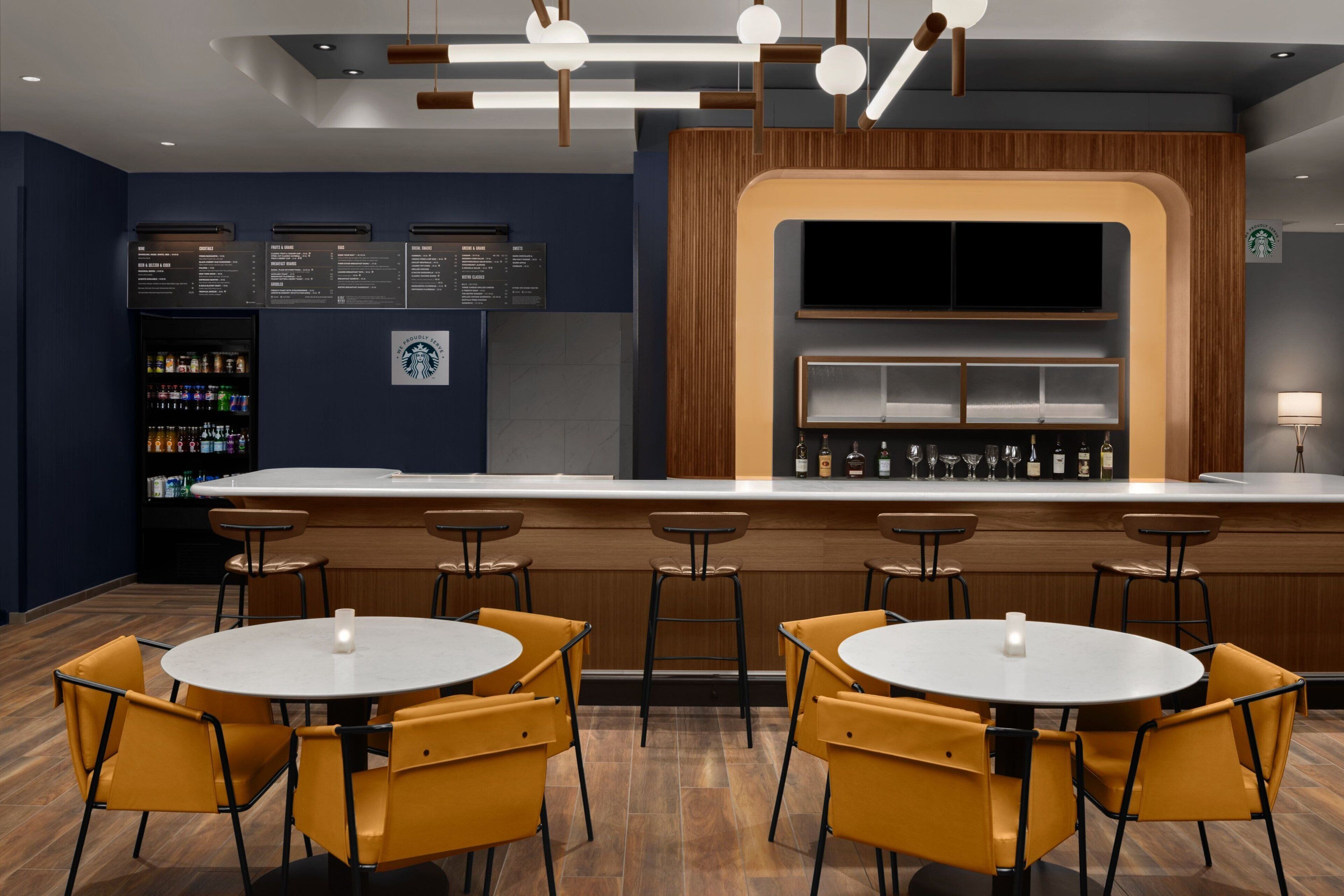 Zona de bar moderna con mesas redondas y sillas amarillas, una barra de madera y un panel con la carta.