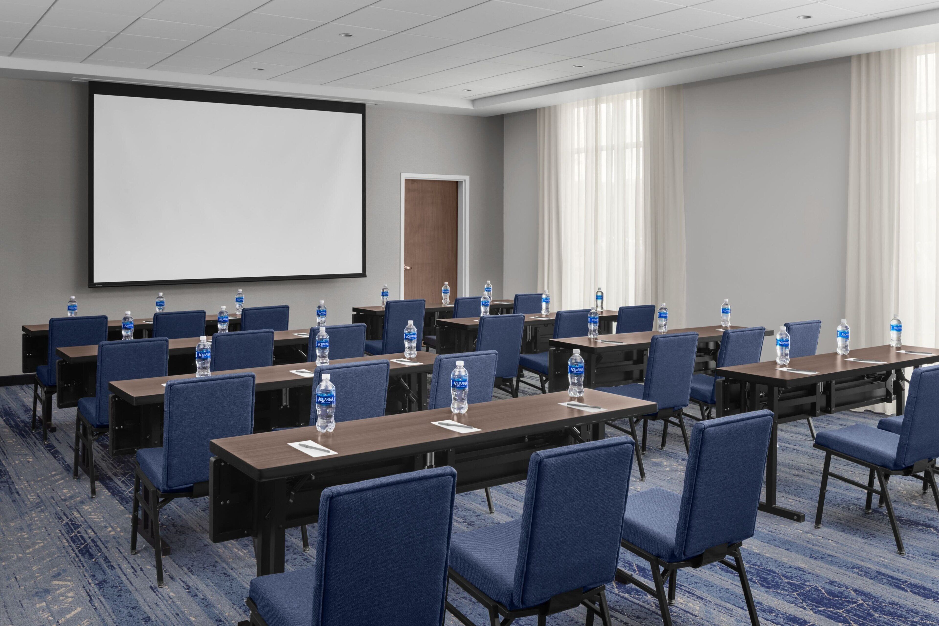 A conference room with rows of tables and chairs, water bottles on each table, a projector screen, and a door in the background.
