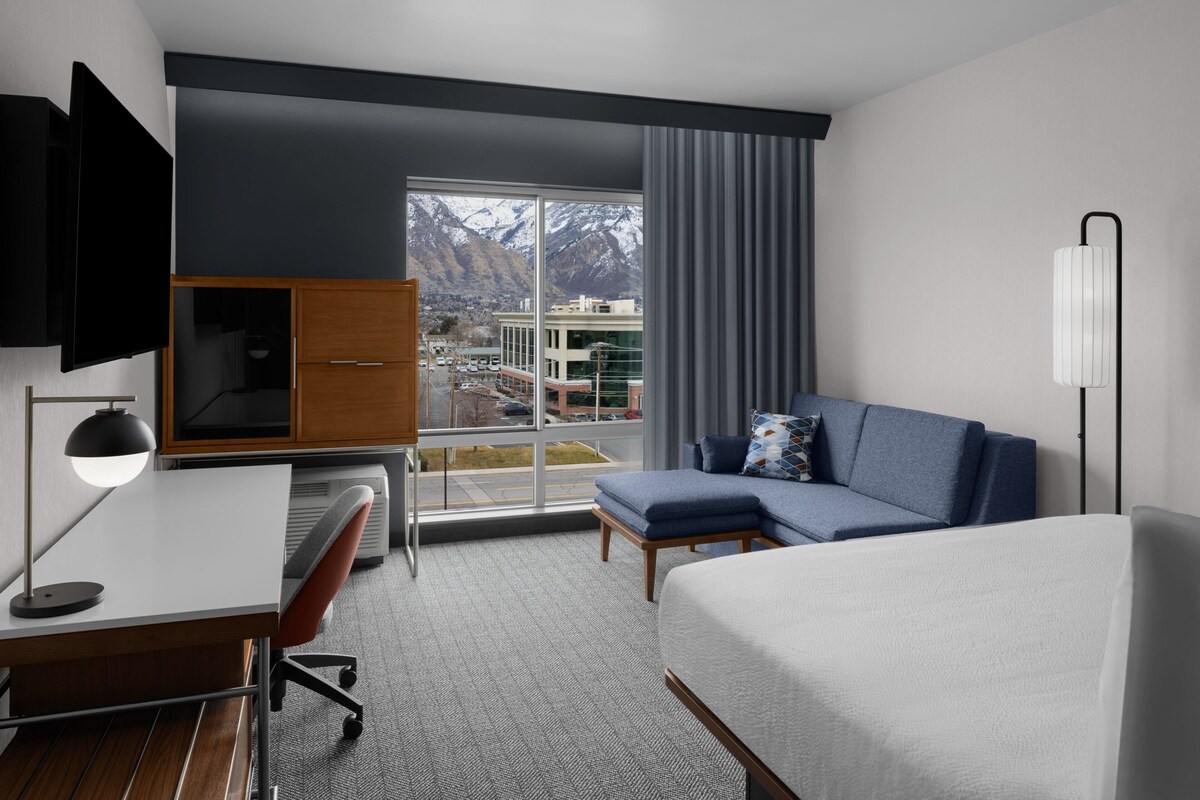 A hotel room with a bed, a desk, a chair, a sofa, a TV, and a large window with a view of mountains and buildings.