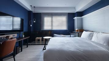 A modern hotel room with a large bed, a desk, a chair, a small table, and a window with blinds.