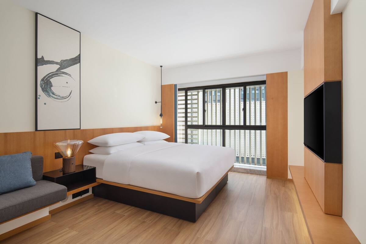 Een moderne slaapkamer met een bed, nachtkastje, lamp, bank, televisie en een groot raam met uitzicht op gebouwen.
