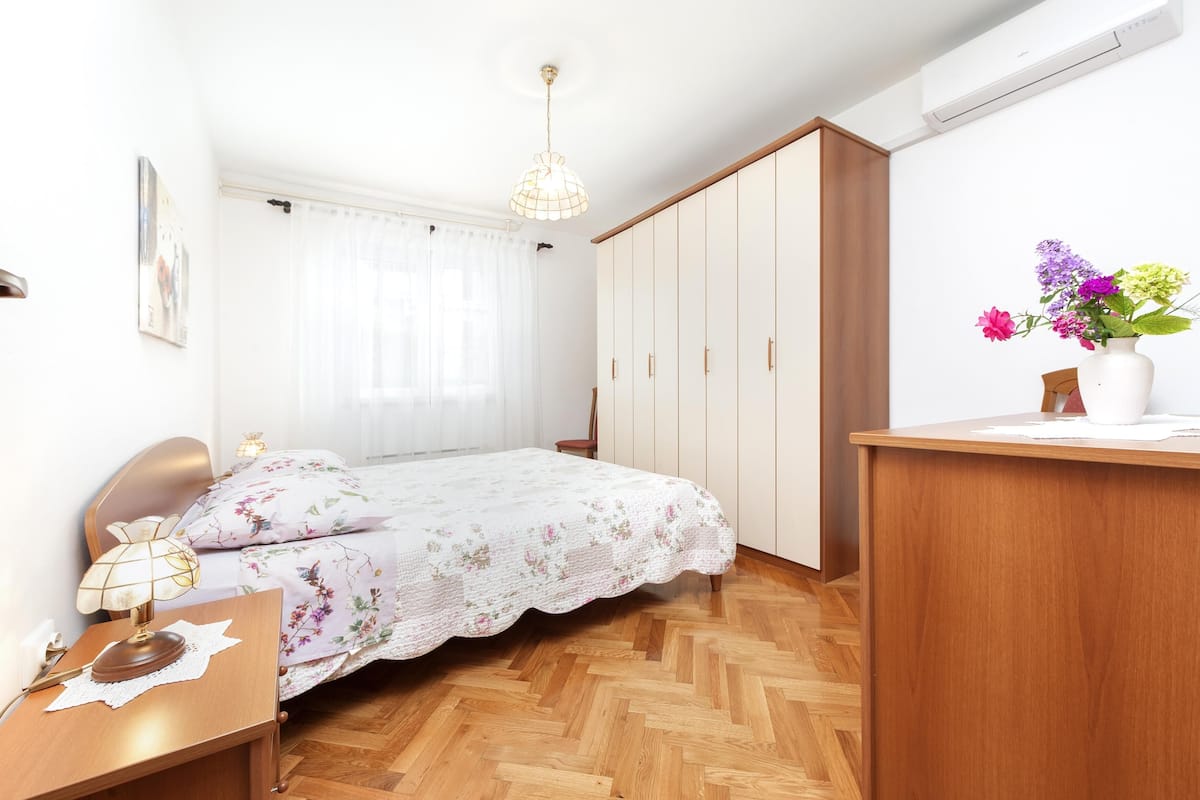 A bedroom with a wooden bed, a bedside table with a lamp, a wardrobe, and a desk with a vase of flowers.