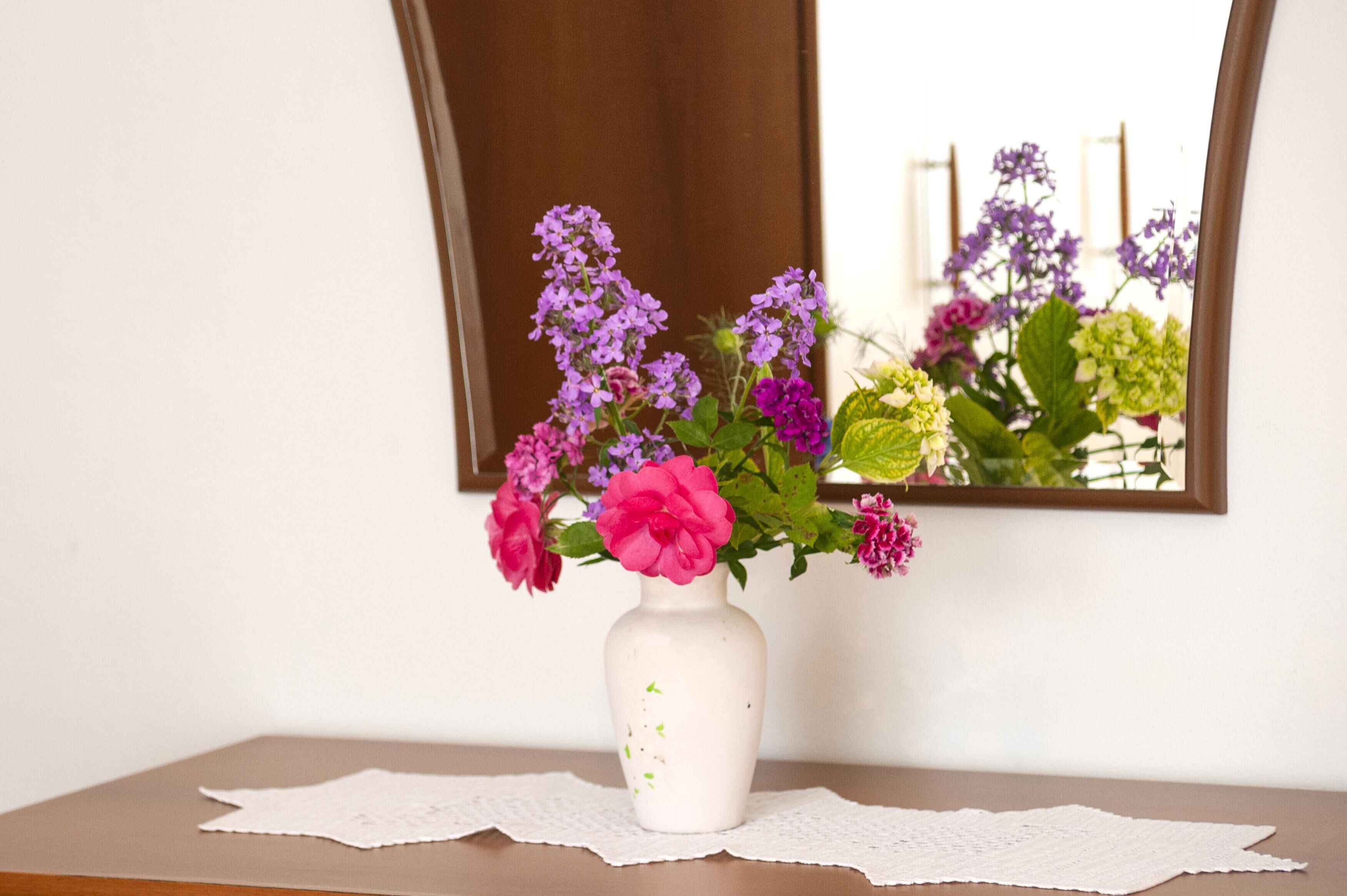 Un vaso bianco con fiori rosa e viola su un tavolo di legno con un sottobicchiere di pizzo.