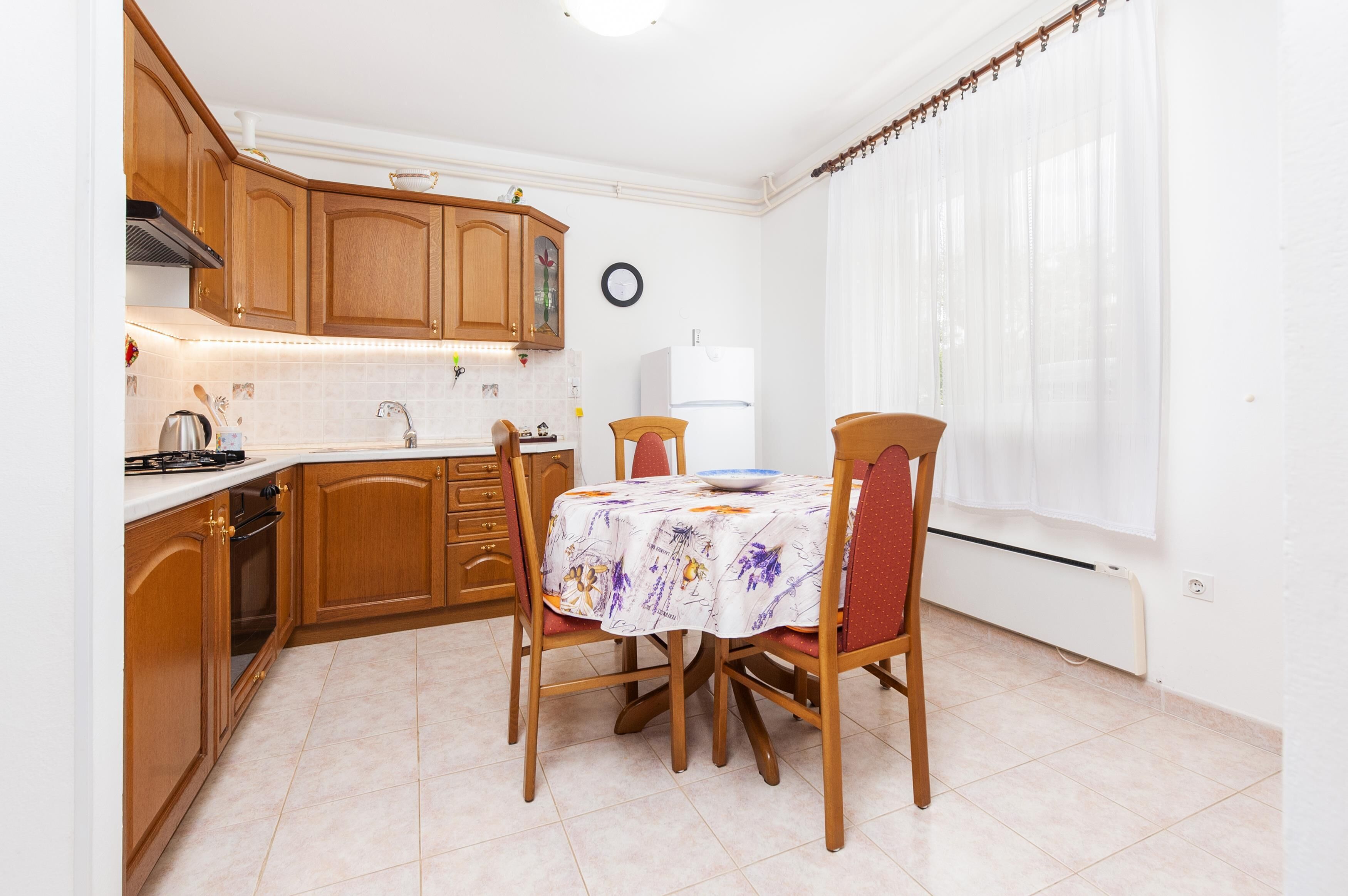 A compact kitchen with wooden cabinets, a dining table with chairs, a refrigerator, and a clock on the wall.