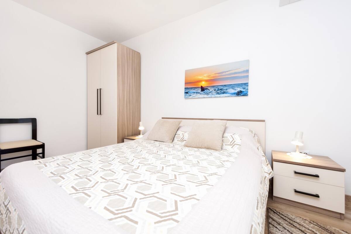 A bedroom with a bed, nightstands, a wardrobe, and a wall-mounted picture of a sunset over the ocean.