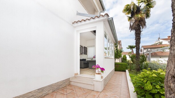 Un bâtiment blanc doté d’un patio carrelé, d’un petit jardin avec un palmier et d’une porte d’entrée donnant sur un espace intérieur.