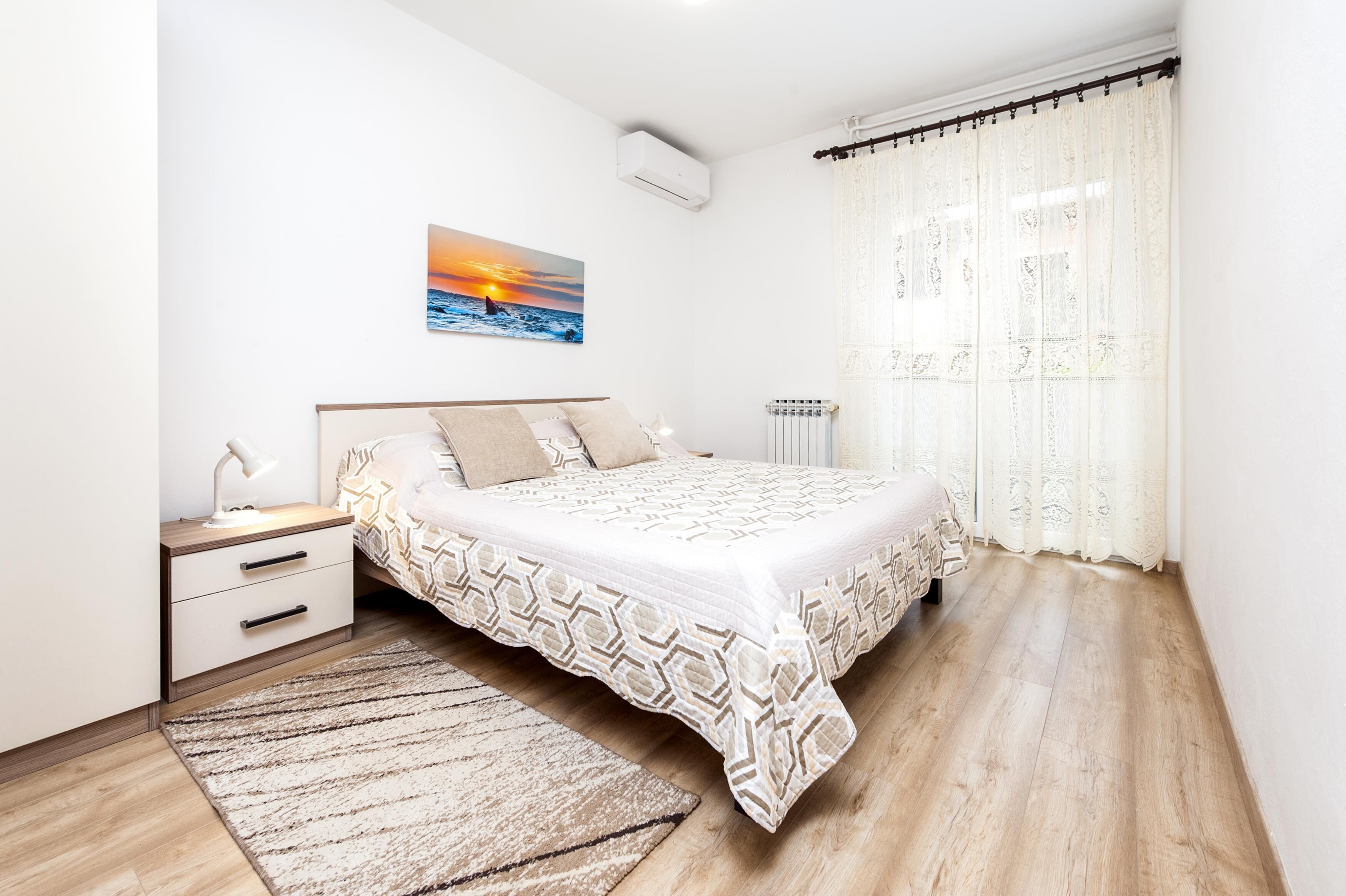 A bedroom with a bed, bedside table, lamp, and a painting of a sunset.