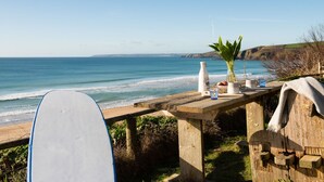Ein Holztisch mit einem Surfbrett, einer Flasche, einer Vase mit grünen Blättern sowie Tassen und einer Kaffeetasse mit Blick auf die Küste.