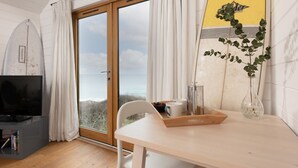 Ein Zimmer mit Holzboden, einem weißen Tisch mit Tablett, einem weißen Stuhl, einem Fernseher, einem Surfbrett und Blick auf das Meer durch eine Glastür.