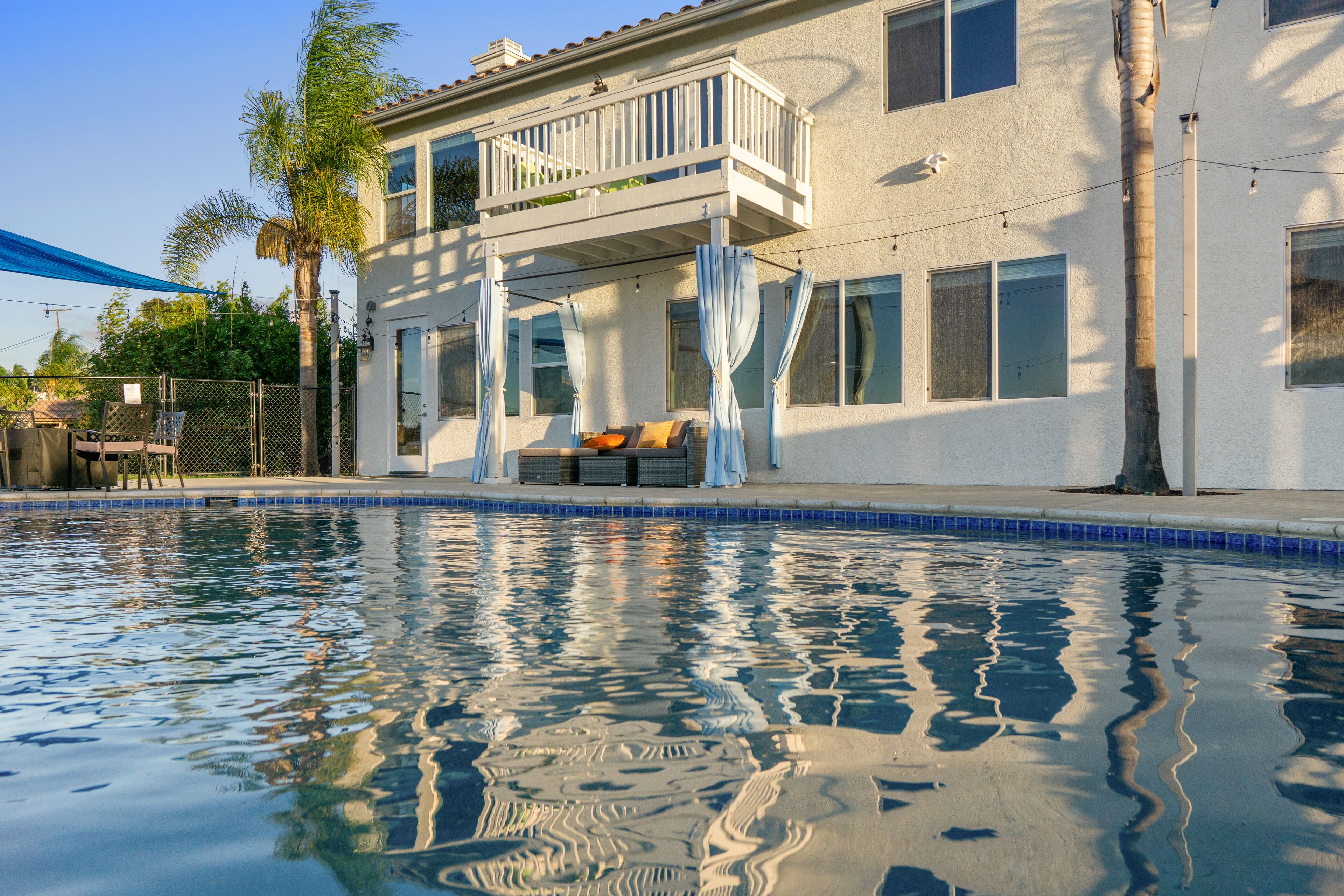A residential pool area with a two-story house, a balcony, and a covered seating area.