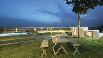 Un espace extérieur aménagé avec une table et des bancs, un arbre, et une vue sur la mer au crépuscule.