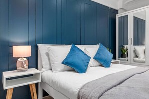Ein Schlafzimmer mit einem weißen Bett, blauen Kissen, einer Nachttischlampe und einem Kleiderschrank.