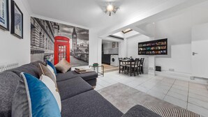 Ein modernes Wohnzimmer mit grauem Sofa, einer Wandmalerei eines roten Telefonhäuschens und einem Flachbildfernseher. Im Hintergrund ist eine Küche zu sehen.